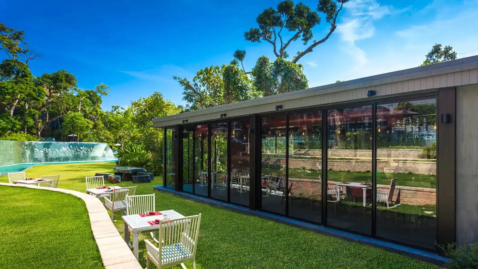 The glass pavilion of Baba Hot Box on a lawn, with garden tables and a waterfall pool behind