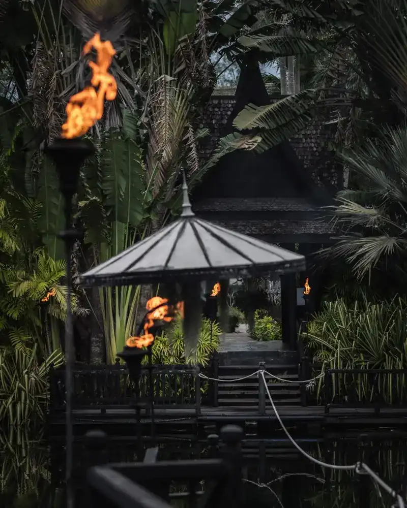 Torches burning by a dark pavilion, with the wooden house behind among palms