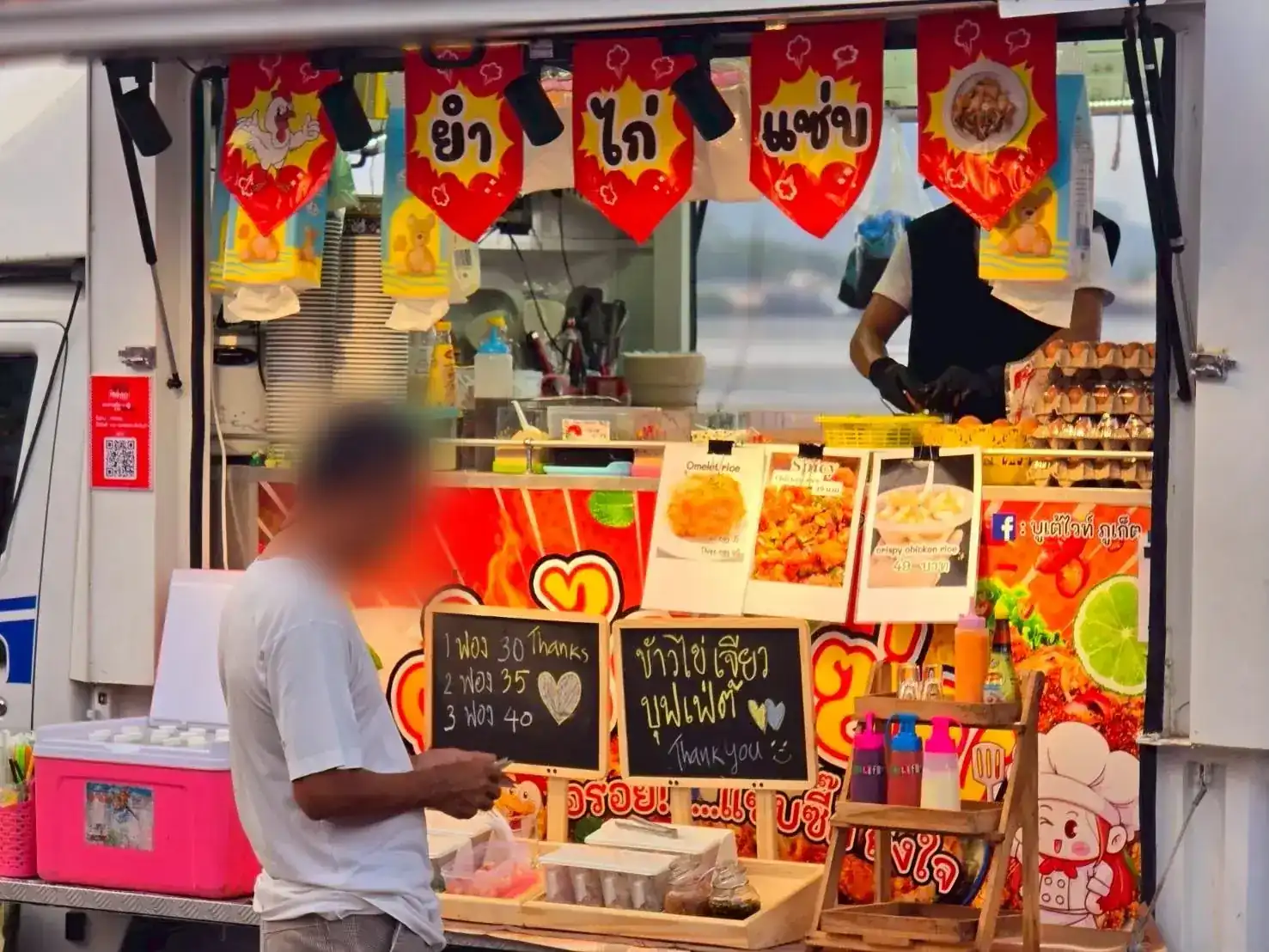 The truck's open hatch: red flags reading ยำ ไก่ แซ่บ, chalkboards for omelette rice, the cook at work inside and a customer at the counter