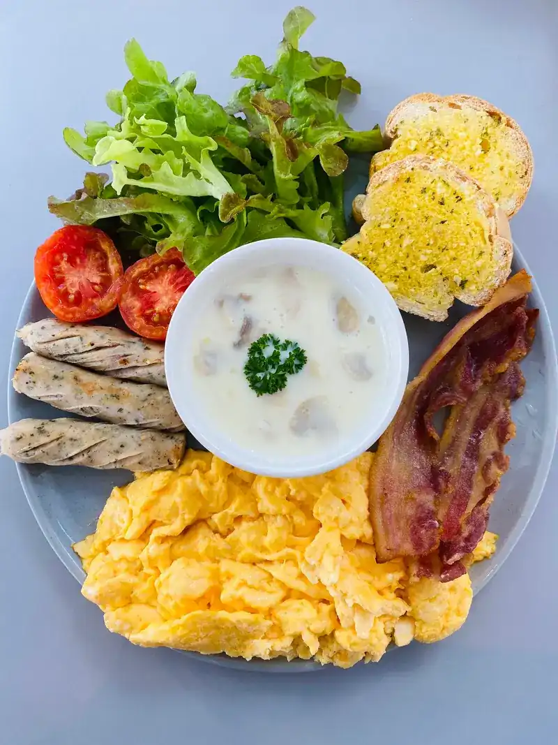 A breakfast plate from above: scrambled eggs, bacon, grilled sausages, garlic bread, tomatoes, salad and a bowl of creamy soup.