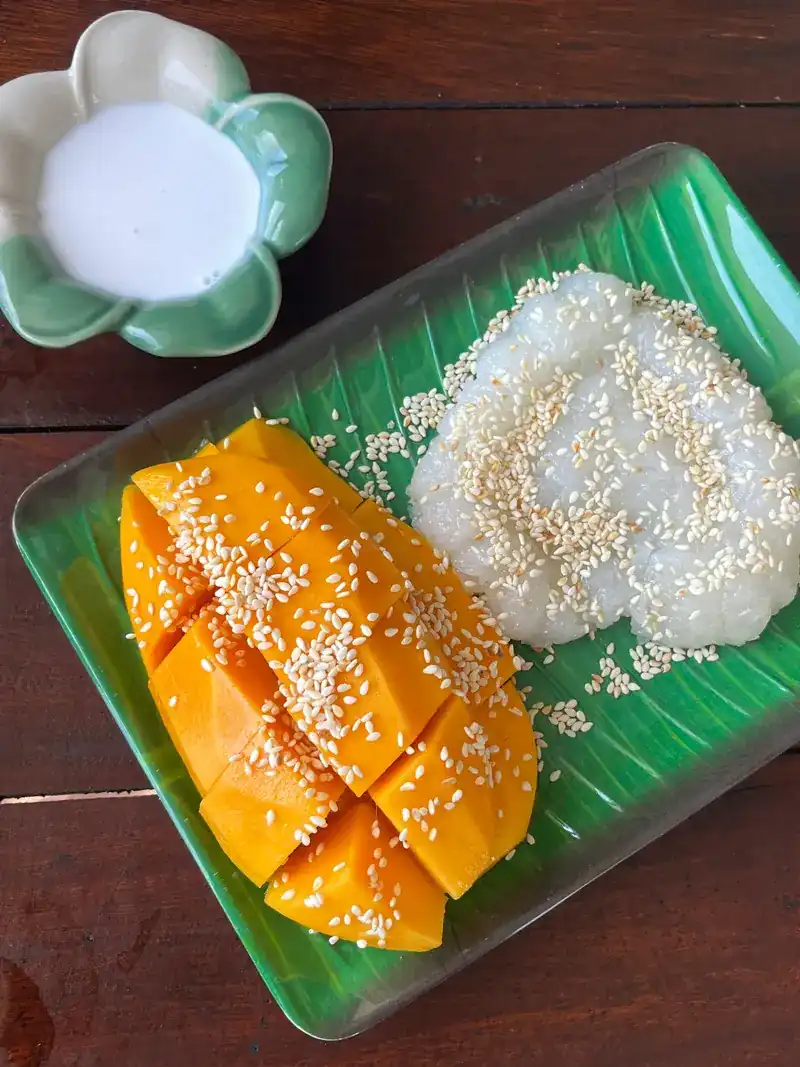 Ripe mango slices and sticky rice with sesame on a green leaf-shaped plate, with coconut cream in a flower-shaped bowl