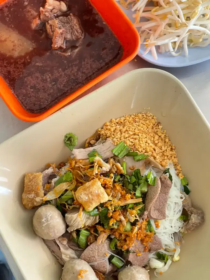 Dry noodles topped with ground peanuts, crispy pork, meatballs and spring onions, with a bowl of soup and bean sprouts beside them