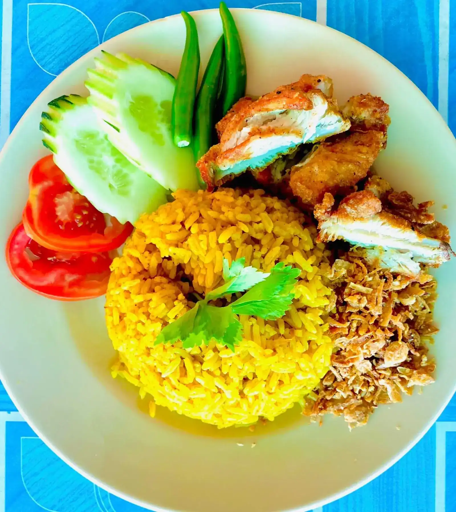 Yellow rice with fried chicken, a heap of fried shallots, cucumber, tomato and green chillies, on a blue tablecloth