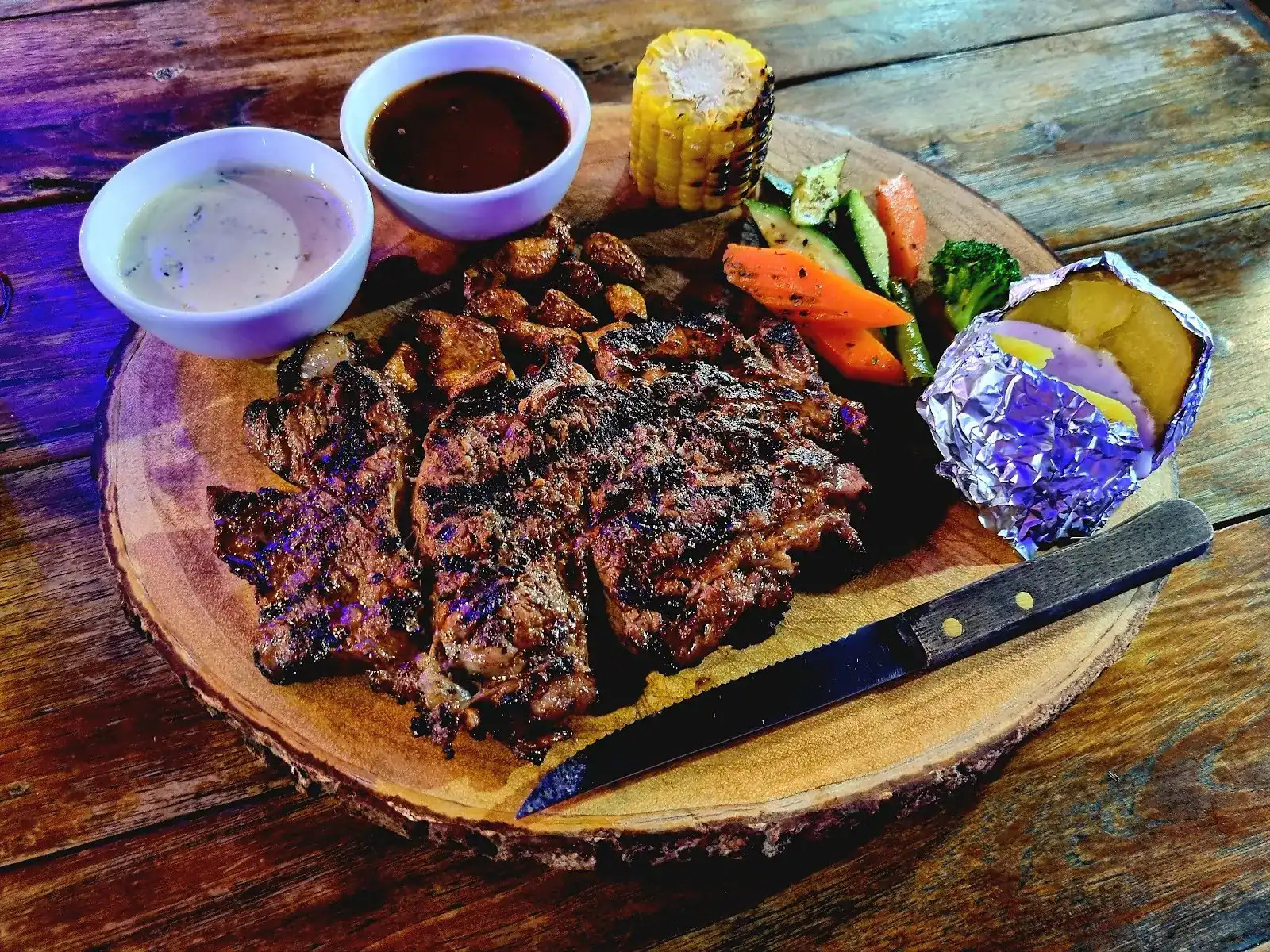 Grilled steak on a wooden board with two sauces, grilled corn, vegetables and a baked potato