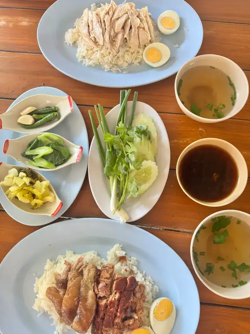 Chicken rice and a plate of pork on rice with boiled egg, with bowls of soup, dark sauce and fresh greens on a wooden table