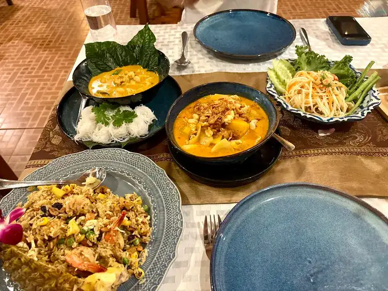 A table of dishes: two orange curries, rice noodles with coriander, papaya salad with long beans, and pineapple fried rice with prawns
