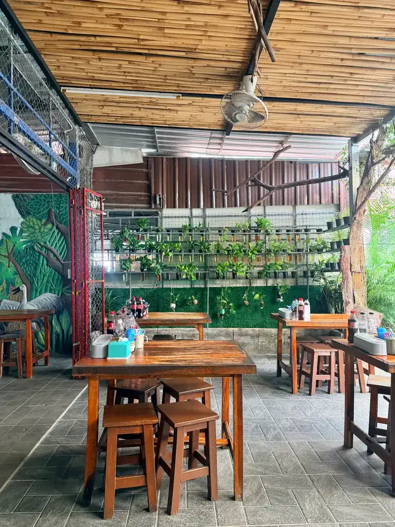 High wooden tables under a bamboo ceiling, beside a wall of potted plants