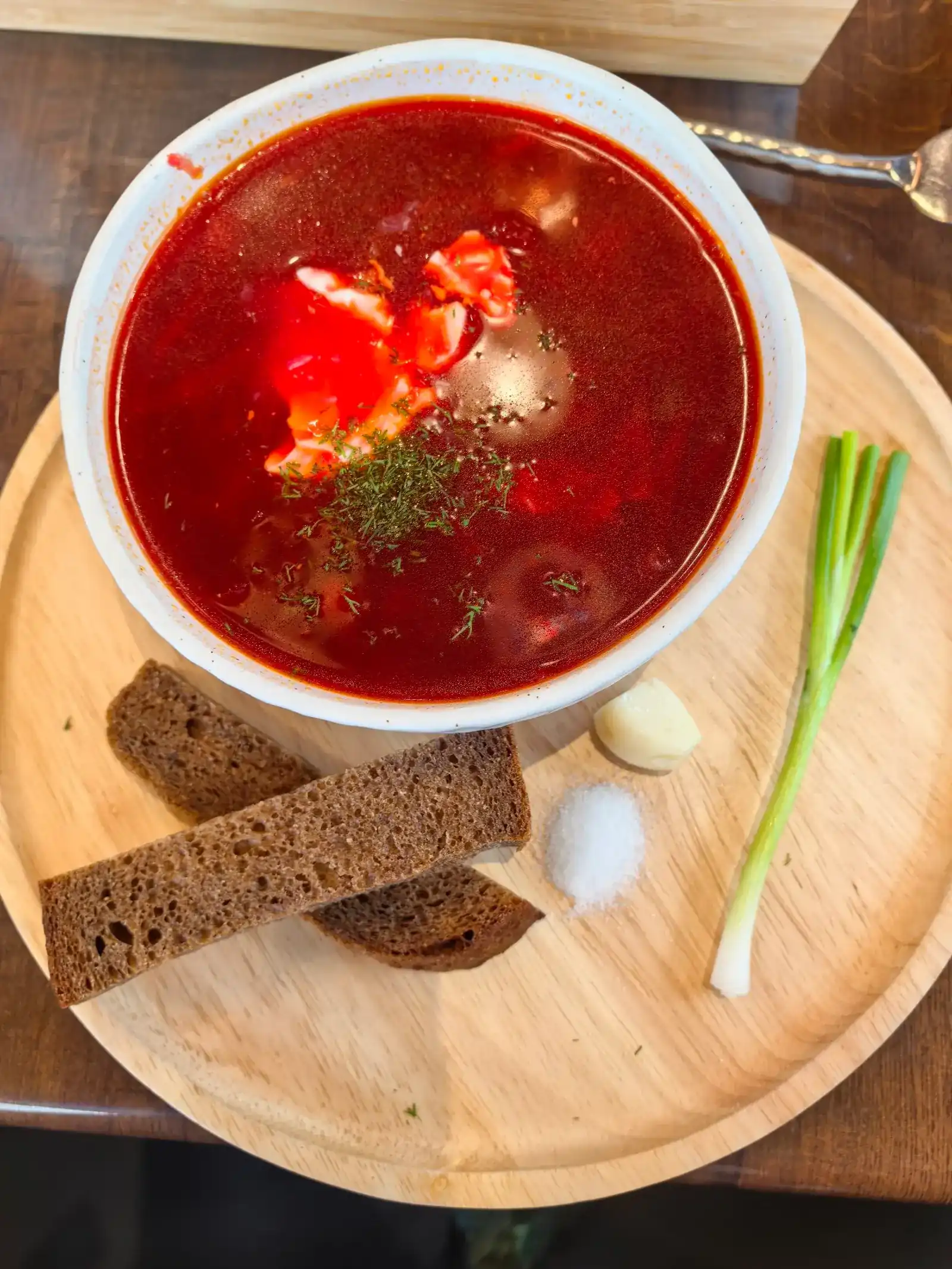 Borscht topped with dill, served with rye bread, garlic, salt and a spring onion on a wooden board