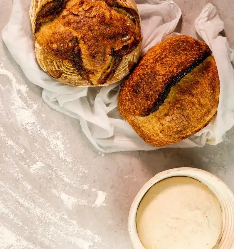 Two sourdough loaves on a linen cloth beside a proofing basket of dough