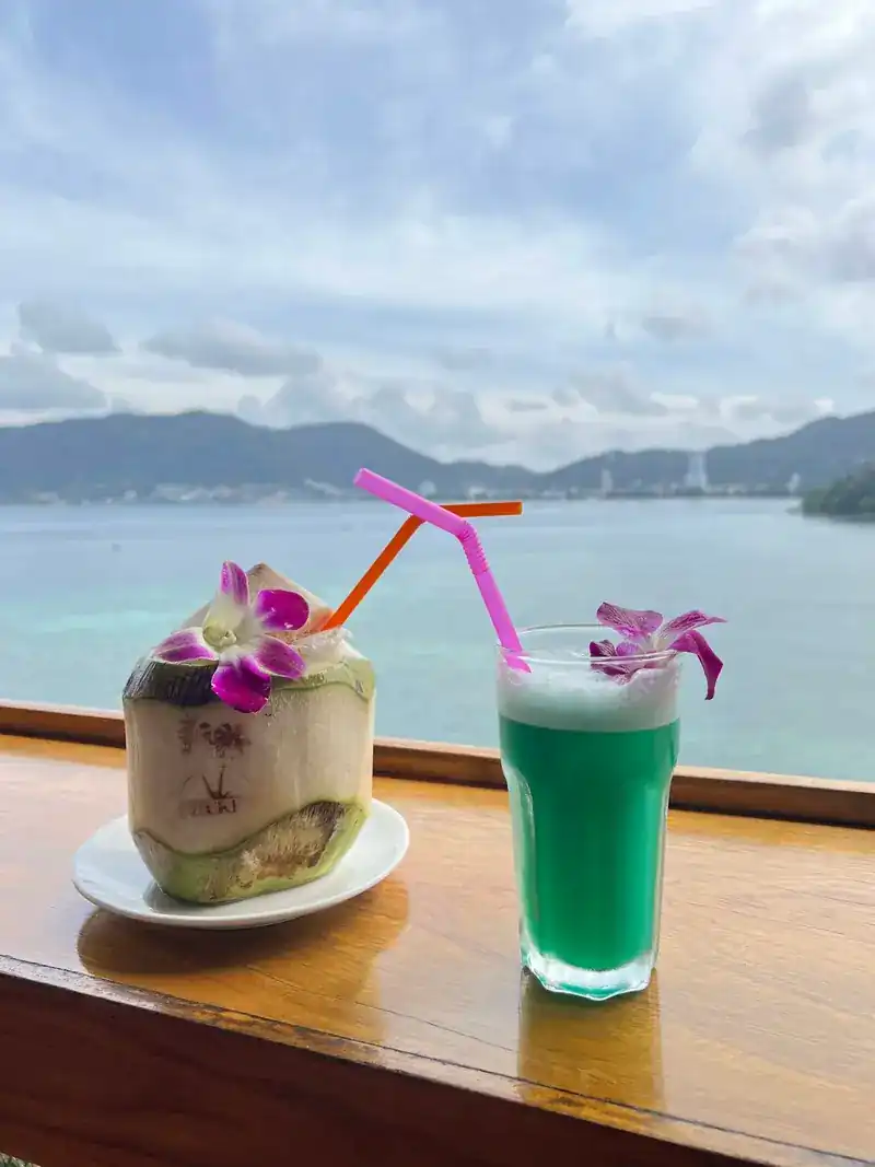 A fresh coconut and a green drink, both with purple orchids, on the railing above Patong Bay