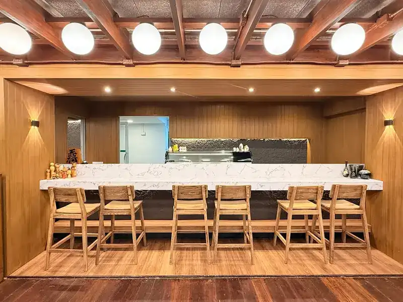 A long white counter with tall wooden stools under round white lanterns