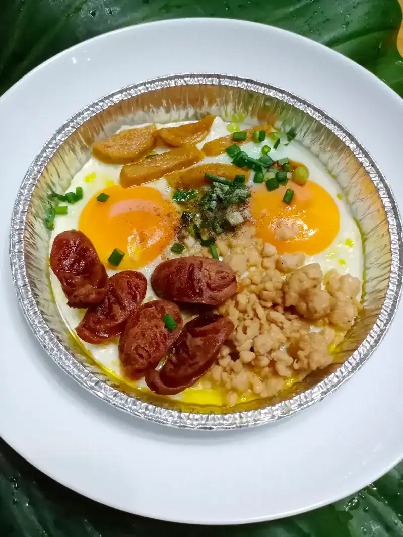 Two eggs cooked in a foil tray with sausage slices, minced meat and spring onion