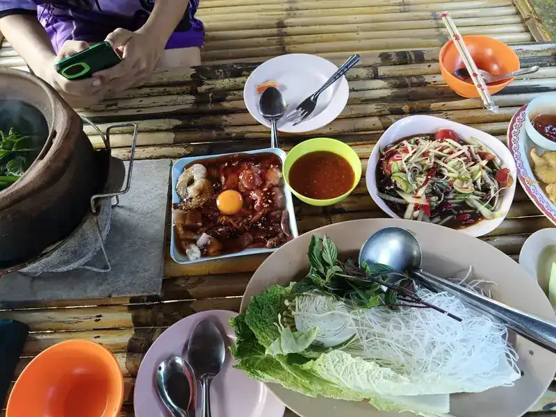 A jim jum table: clay pot of broth, a platter of raw prawns, squid and meat with an egg yolk, vegetables, herbs and noodles, som tam and dipping sauce