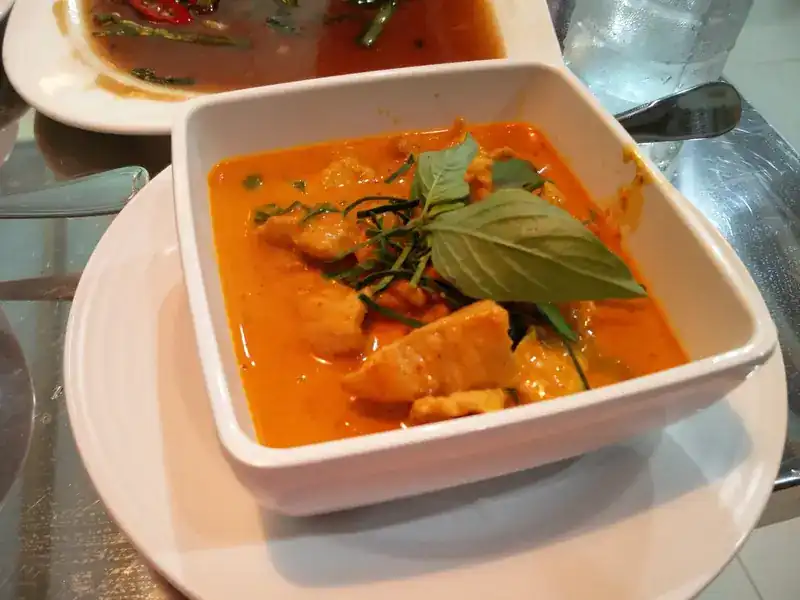 A square white bowl of red curry topped with a basil leaf