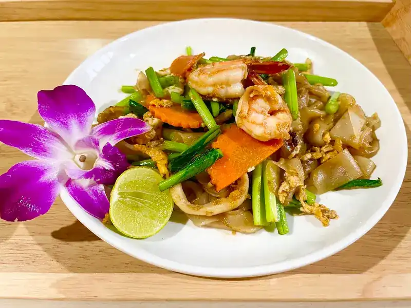Stir-fried wide rice noodles with prawns, squid, carrot and greens, lime and an orchid on the side