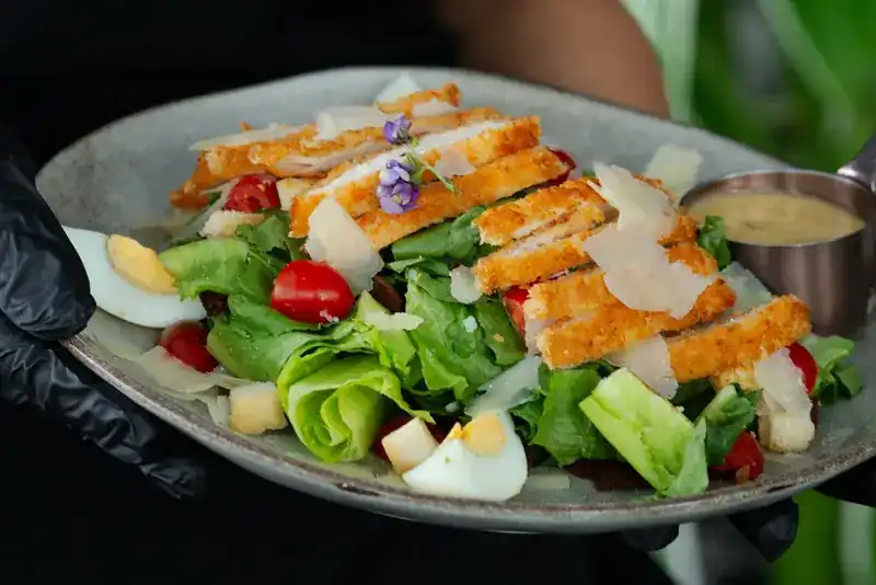 A salad with crispy chicken, egg, cherry tomatoes and shaved cheese, carried to the table