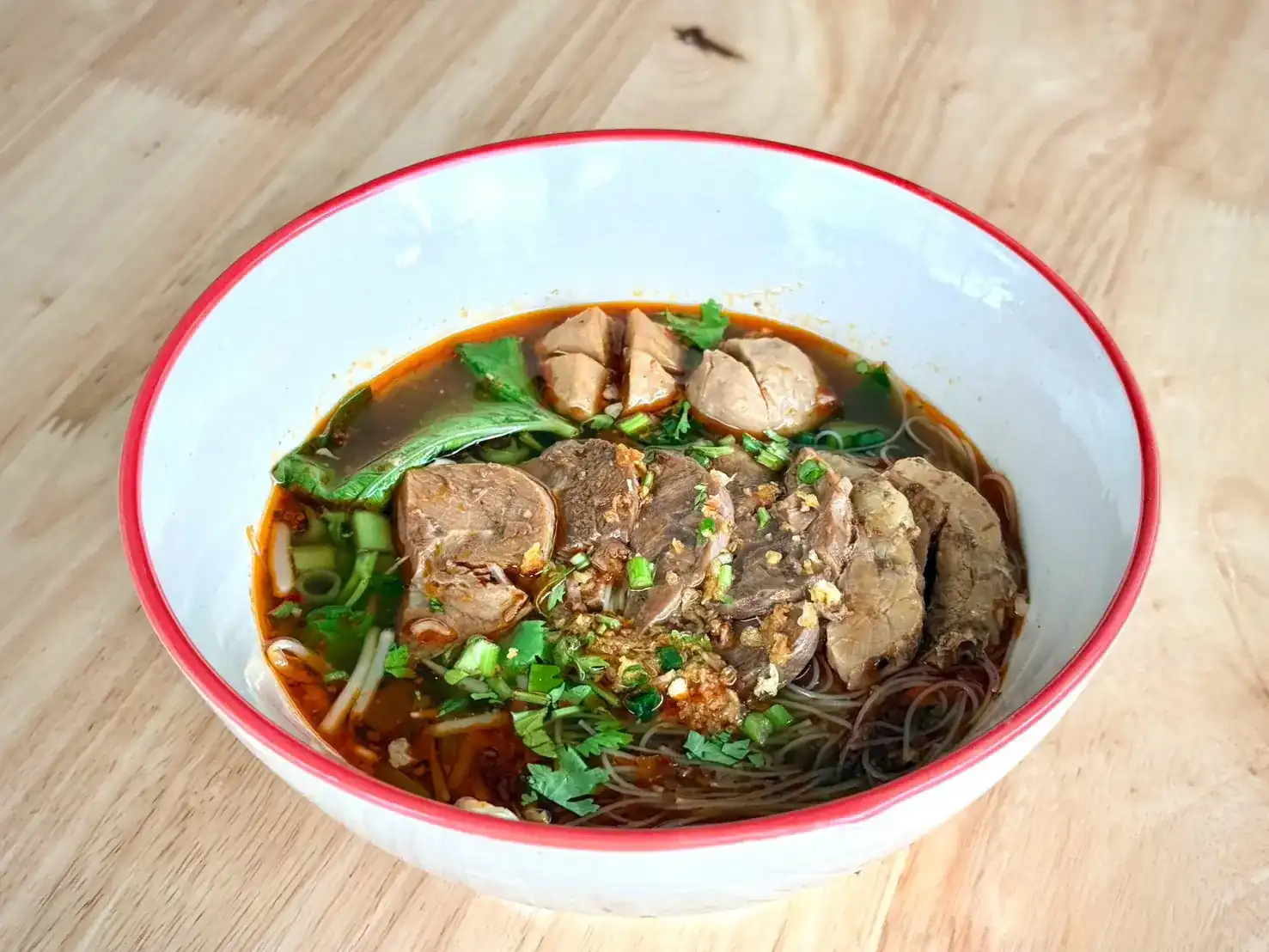 Boat noodles in a red-rimmed bowl: thin rice noodles, sliced meat, meatballs and fresh herbs in a dark broth