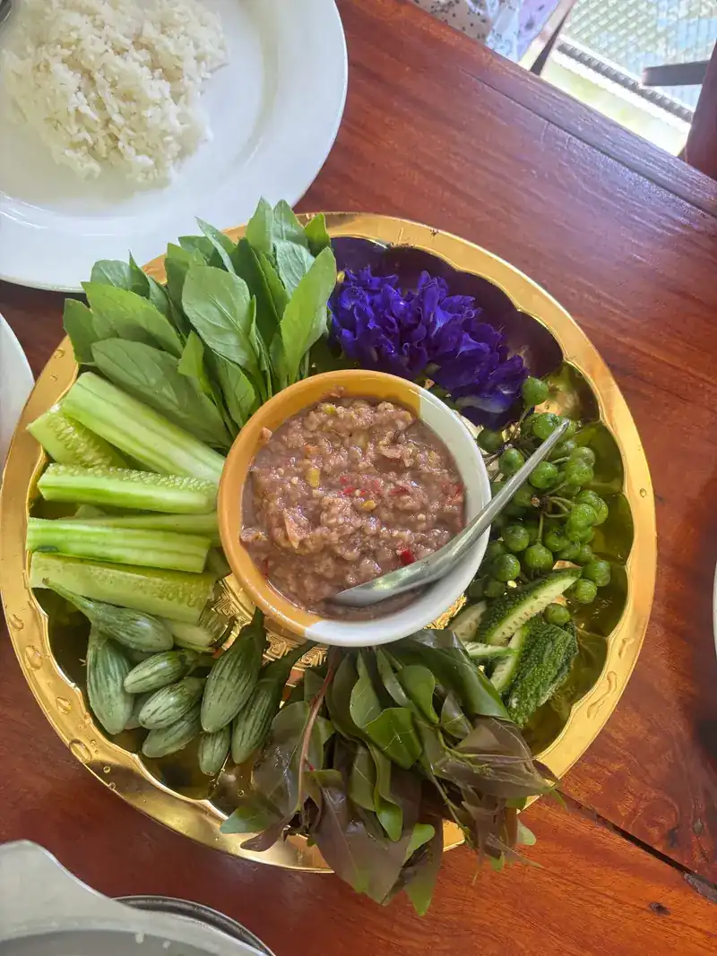 A bowl of nam prik in the centre of a tray of fresh vegetables, leaves and butterfly-pea flowers