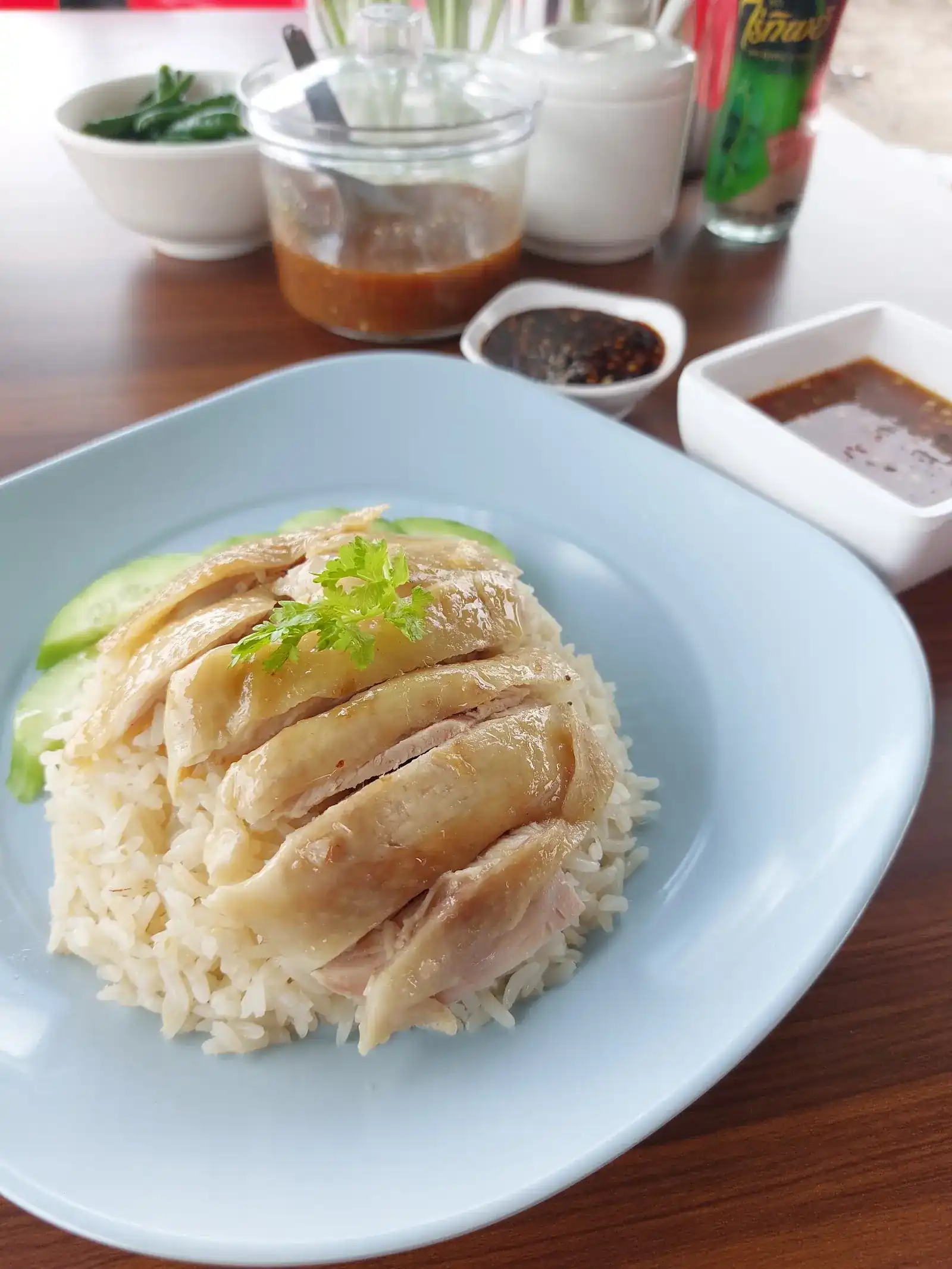 Boiled chicken sliced over chicken rice on a pale blue plate, with cucumber and three sauces behind