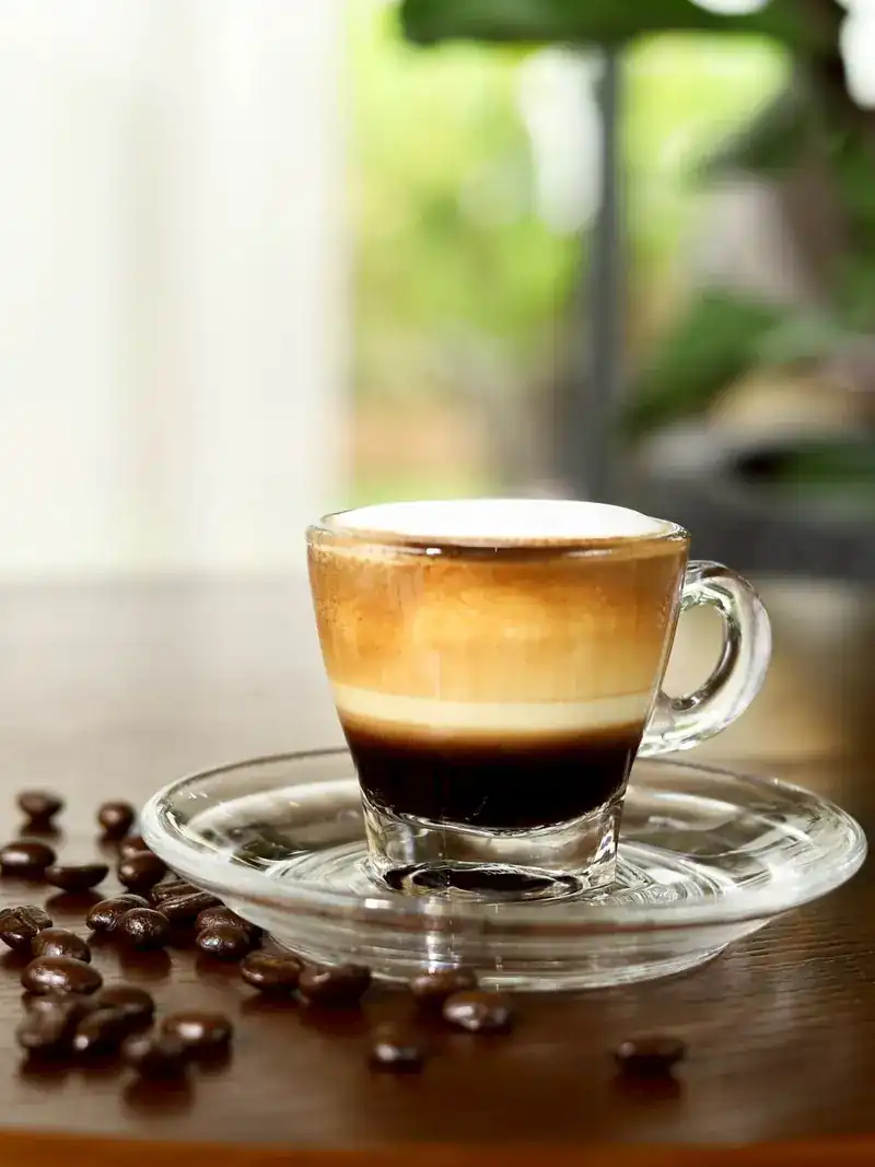 A macchiato in a glass cup, layers of espresso and milk, coffee beans on the table