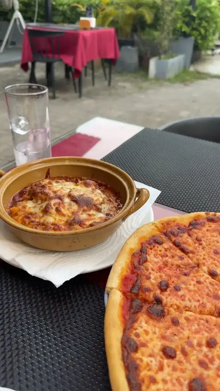 Lasagne in a clay dish and a pizza on a table outside, red tablecloths behind