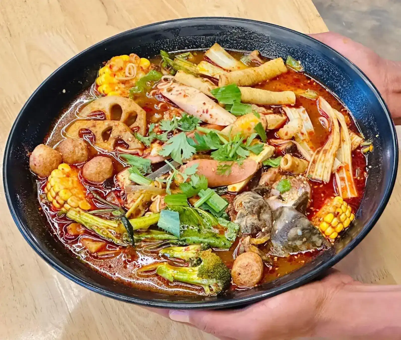 A big bowl of malatang held in two hands: lotus root, sweetcorn, fish balls, tofu skin, broccoli and coriander in red mala soup