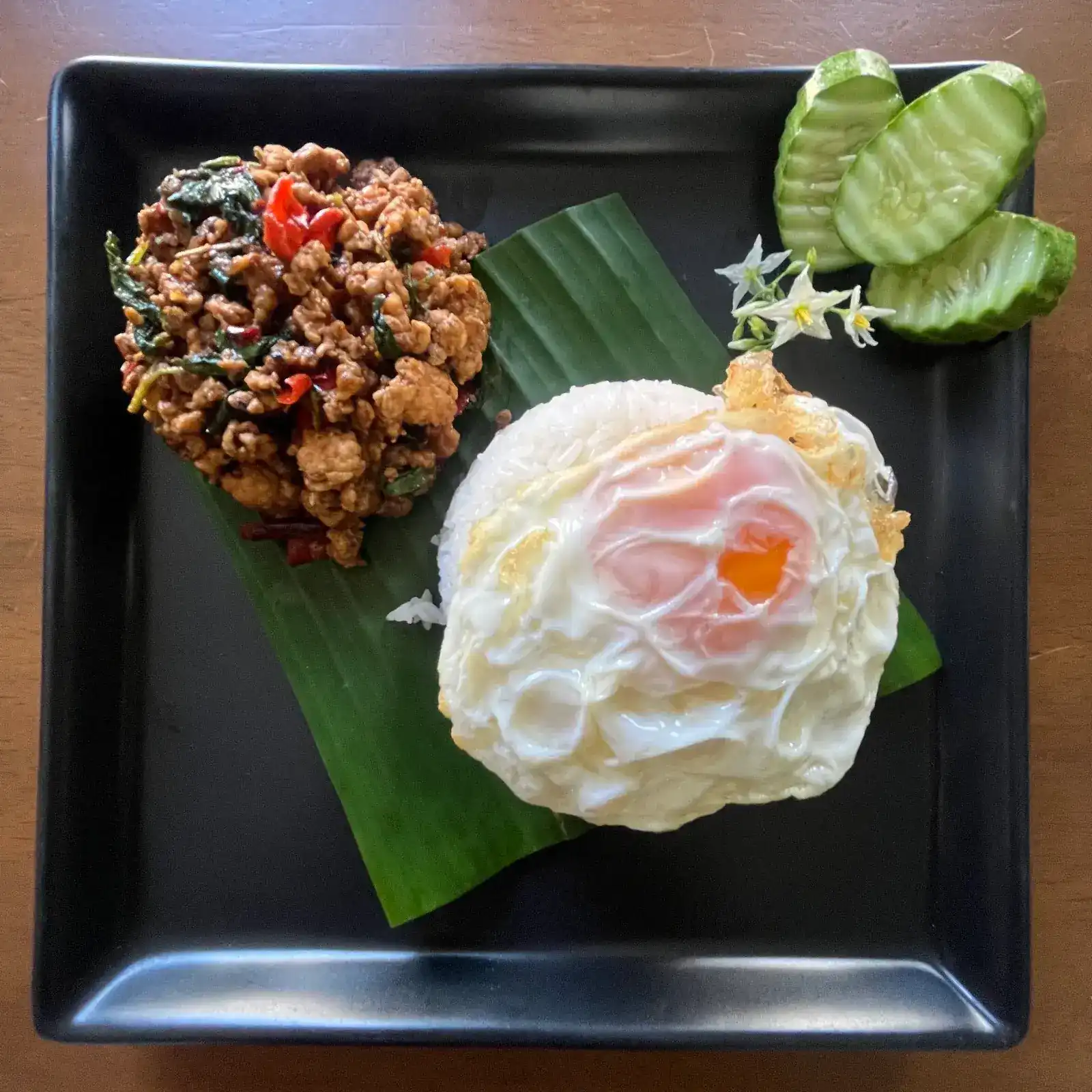 Holy basil stir-fry with rice and a fried egg on a banana leaf, cucumber on the side