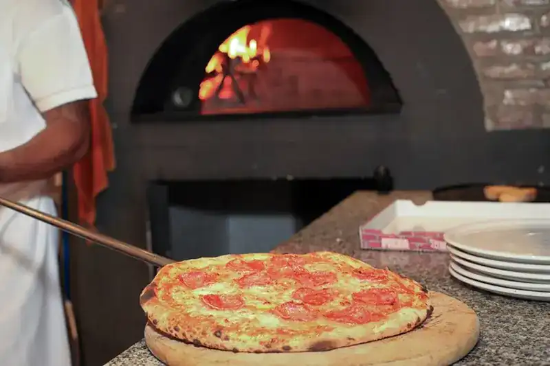 A pizza on a wooden peel in front of the wood oven, the fire burning inside