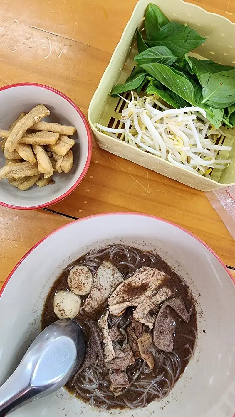 A bowl of boat noodles with a basket of basil and bean sprouts and a bowl of crispy pork
