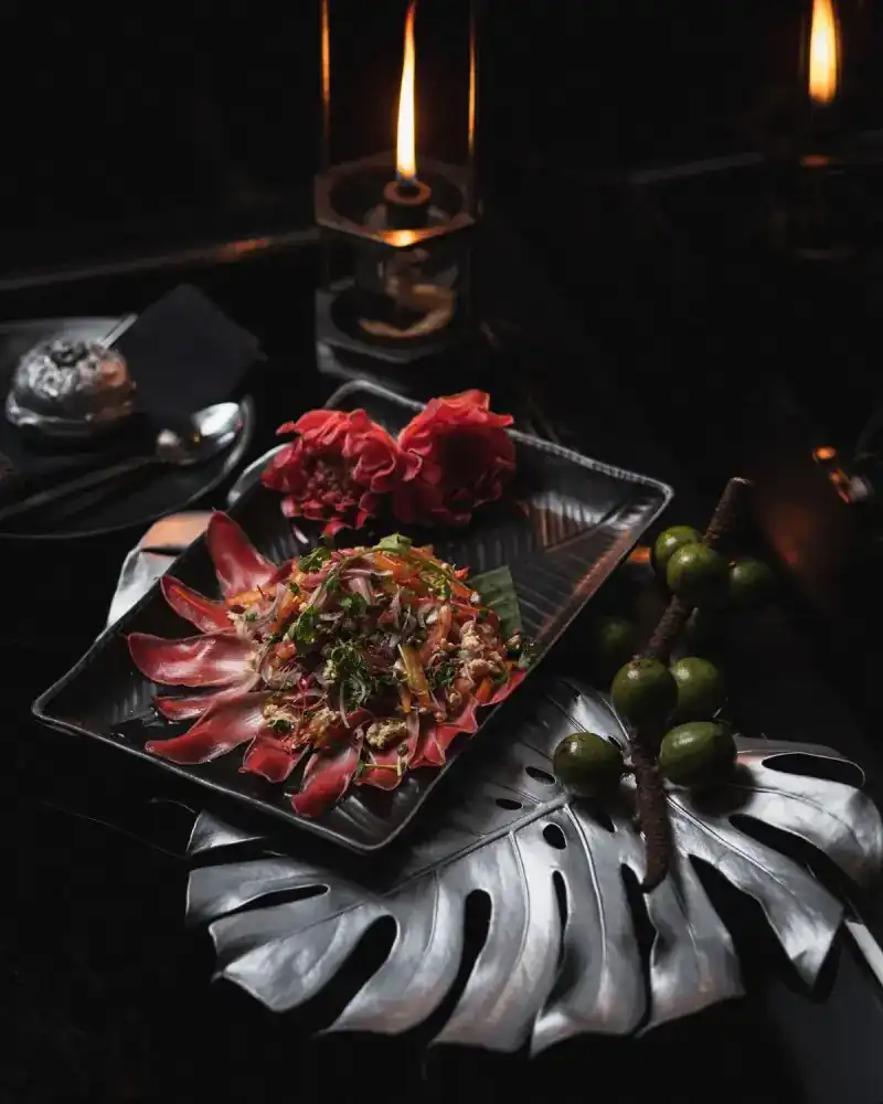 A dressed salad on a black platter with red flowers, on a silver leaf beside a candle lamp