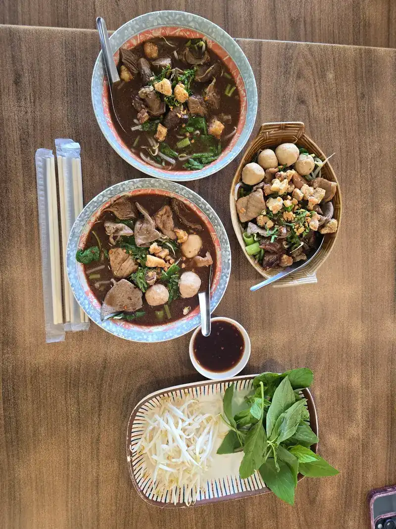 Seen from above: two bowls of boat noodles, a basket of meatballs, bean sprouts, basil and chopsticks