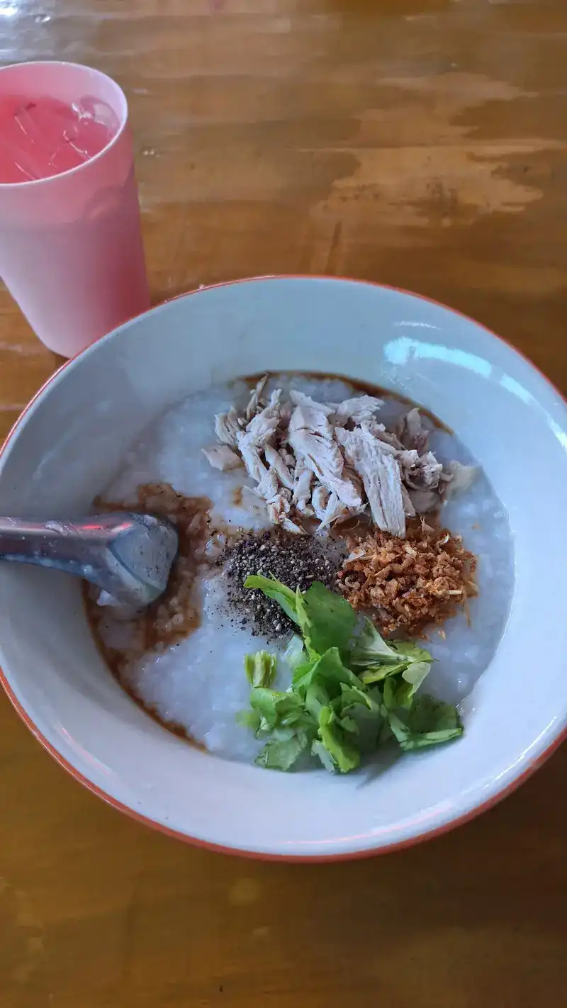 A bowl of rice soup topped with shredded chicken, pepper, fried garlic and fresh herbs, a pink drink beside it