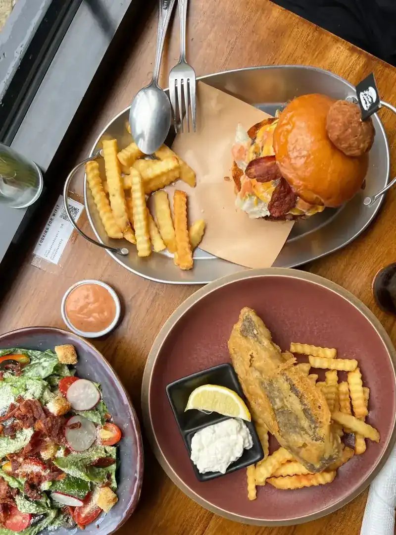 A burger with crinkle-cut fries, fish and chips with tartar sauce, and a salad, on a wooden table