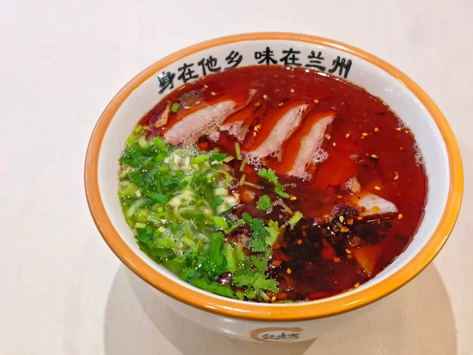 A bowl of Lanzhou beef noodles in red chili-oil broth with sliced beef, spring onion and coriander; the rim reads 身在他乡 味在兰州