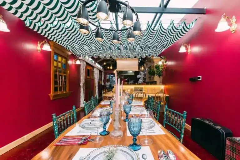 A long wooden table set with blue pressed-glass goblets in a raspberry-red room, under a green and white striped canopy