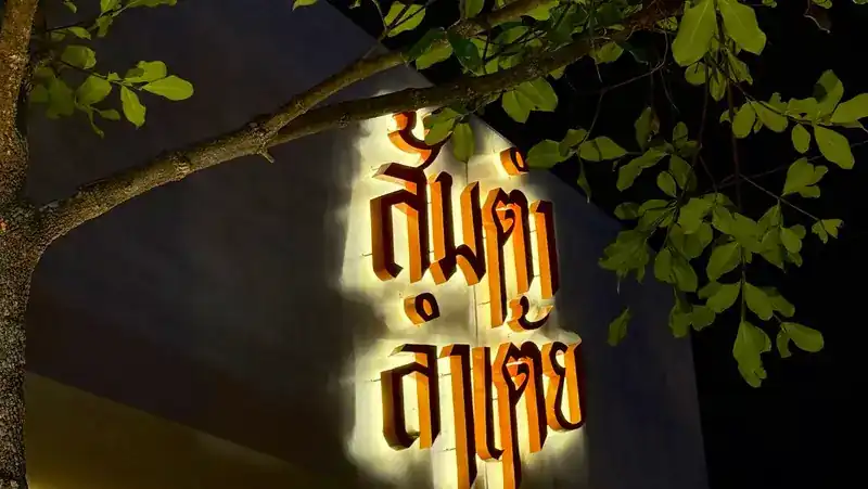 The name ส้มตำลำเต้ย lit up in orange letters on the wall at night, under the branches of a tree