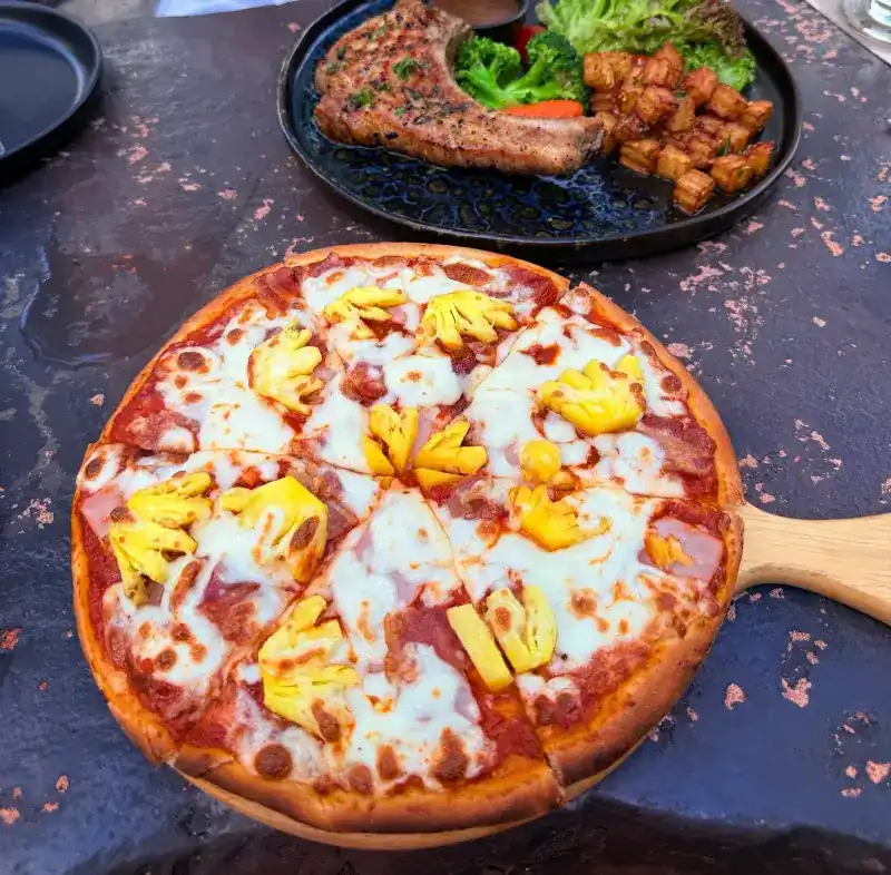Ham and pineapple pizza, with a grilled chop and fried potatoes behind it / พิซซ่าแฮมสับปะรด กับสเต๊กย่างและมันฝรั่งทอด