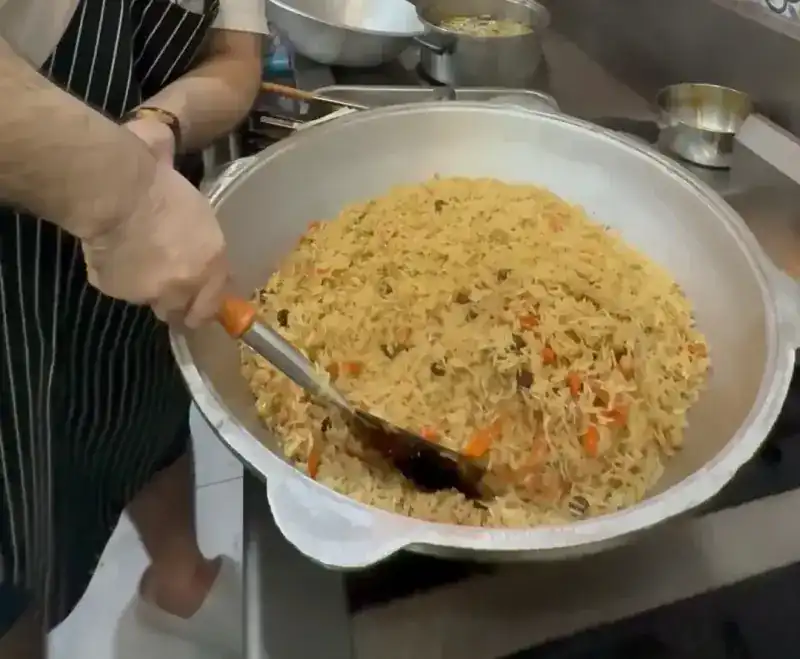 Plov being turned with a long spatula in a wide aluminium kazan in the kitchen