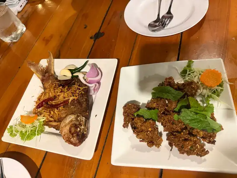 A whole fried fish under crisp shreds beside a plate of fried morsels with fresh herbs, on a wooden table