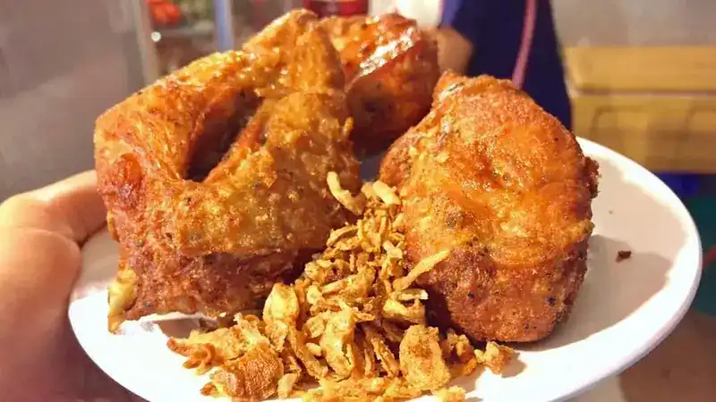 A hand holding a plate of golden fried chicken with crisp fried shallots