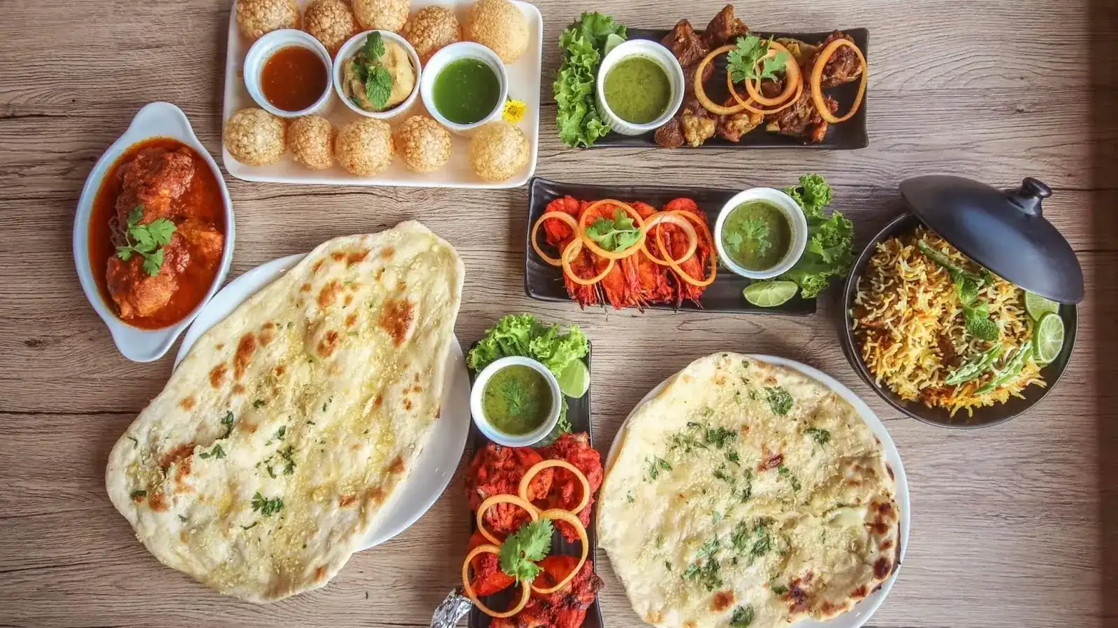 A spread on a wooden table: pani puri with three chutneys, two naan, a curry, tandoori pieces with onion rings and a pot of biryani