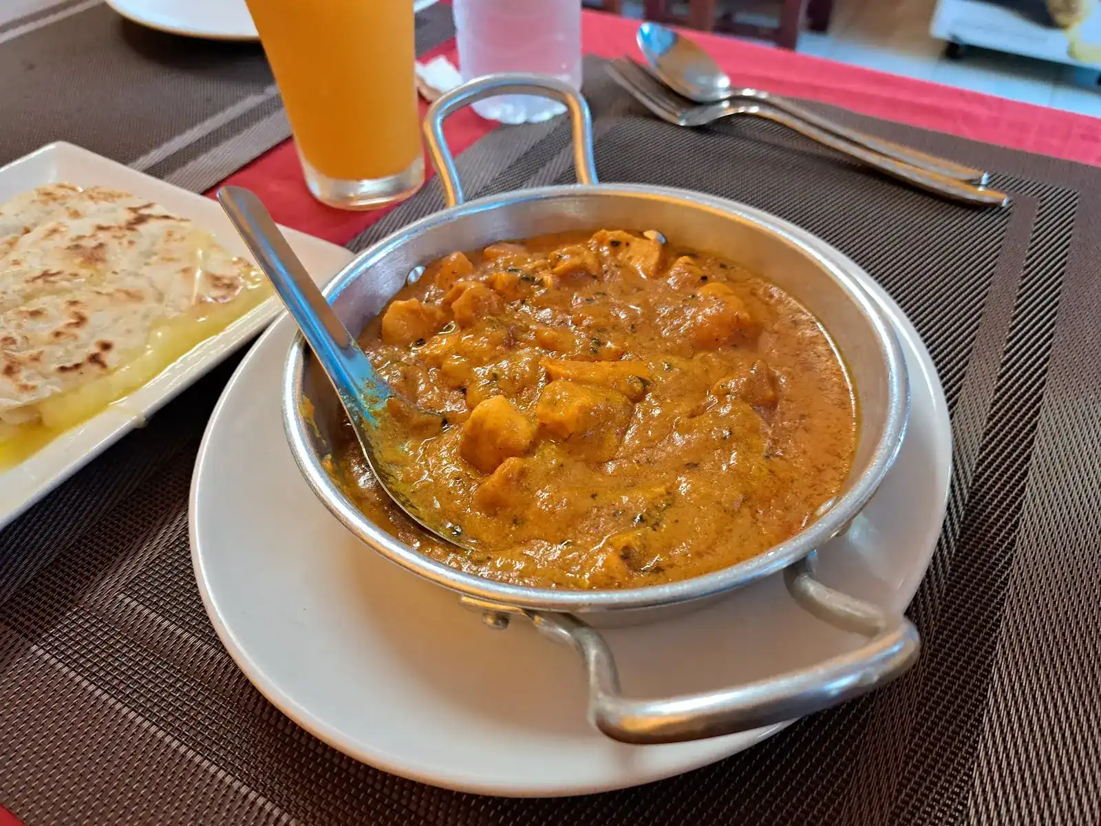 A curry in a steel kadai, with buttered naan and a glass of juice