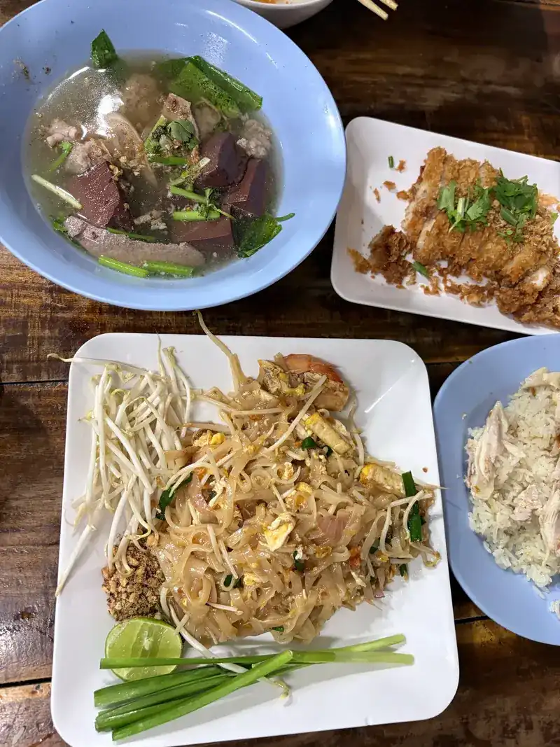 A table seen from above: a bowl of pork soup with pork blood and greens, pad thai with a prawn, fried chicken and chicken rice