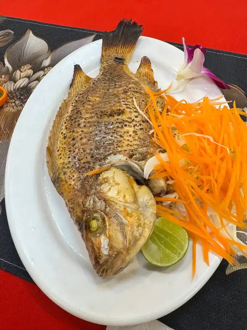 A whole fried fish with shredded carrot, lime and an orchid on a red tablecloth