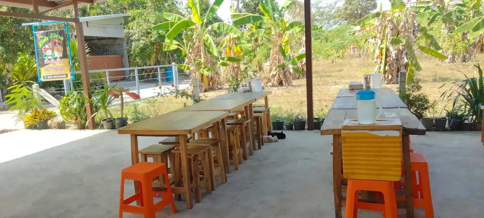 Long wooden tables and orange stools under the open roof, with banana trees beyond