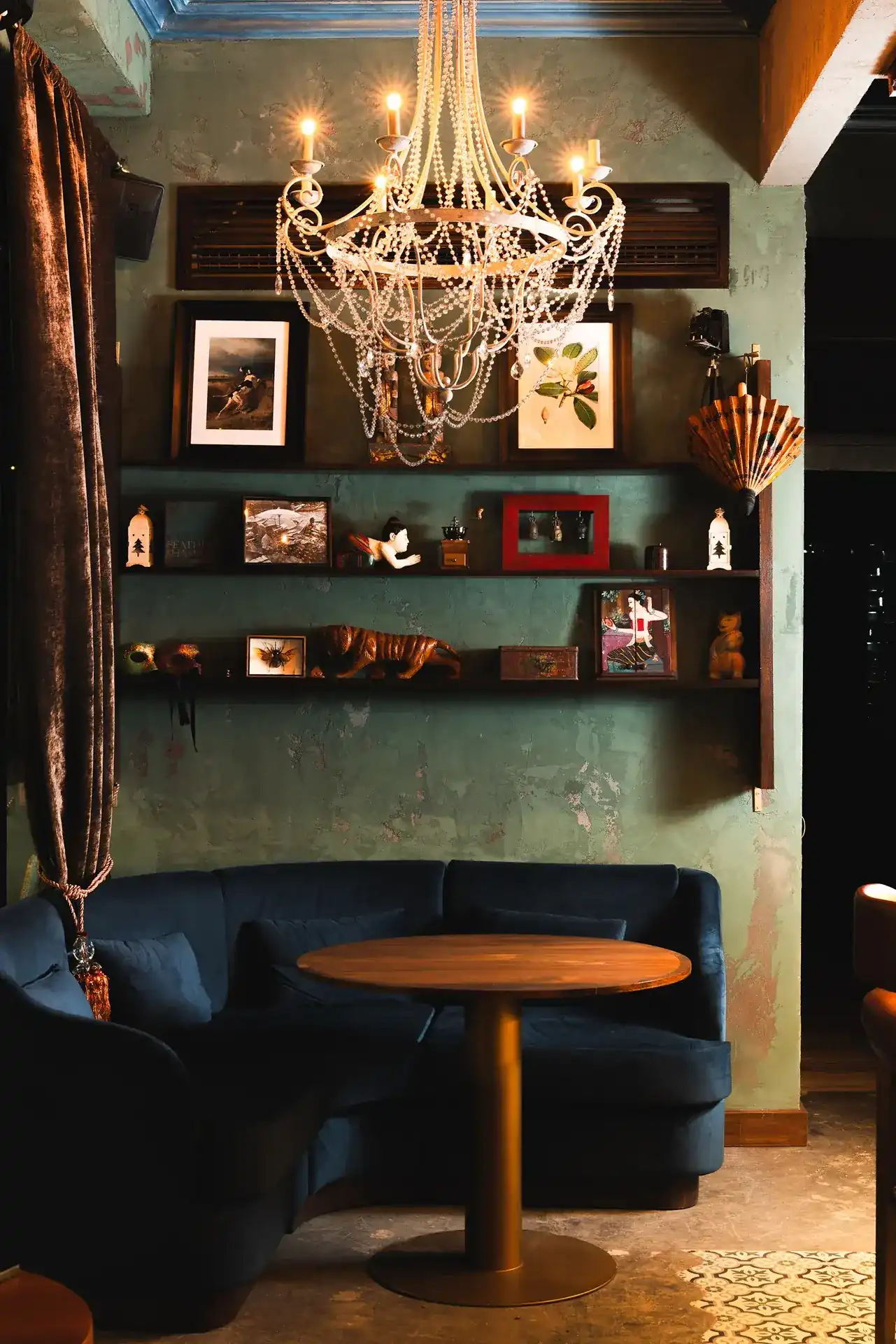 A corner of the room: a navy velvet sofa under a beaded chandelier, with shelves of curios on a green plaster wall