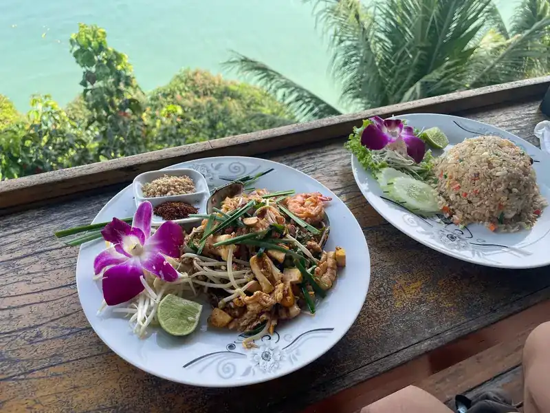 Pad thai with prawns and a plate of fried rice on the wooden railing, trees and sea below