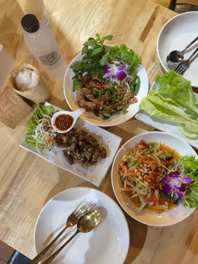 Seen from above on a wooden table: sticky rice in a woven basket, a meat salad with mint and herbs, green papaya salad with peanuts, a fried dish with chilli dip and a bottle of water with the restaurant's own label