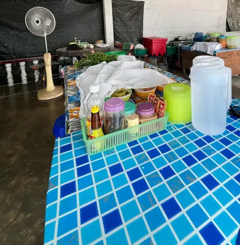 Under the roof: a blue mosaic-tiled table with condiments, water jugs and a standing fan