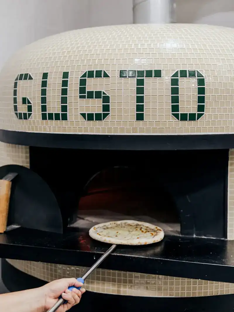A pizza going into the mosaic oven that spells GUSTO in green tiles
